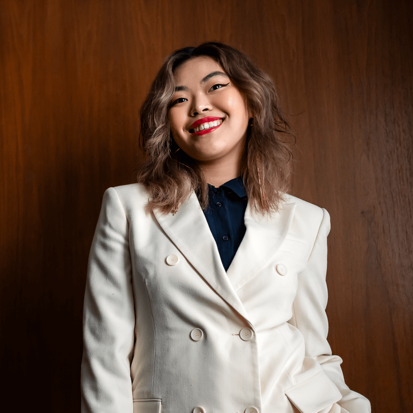 Young person with long brunette hair wearing a white suit smiling against a wood backdrop