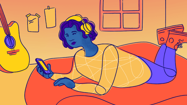 A young person texts on their bed with headphones on, next to a guitar and a window.