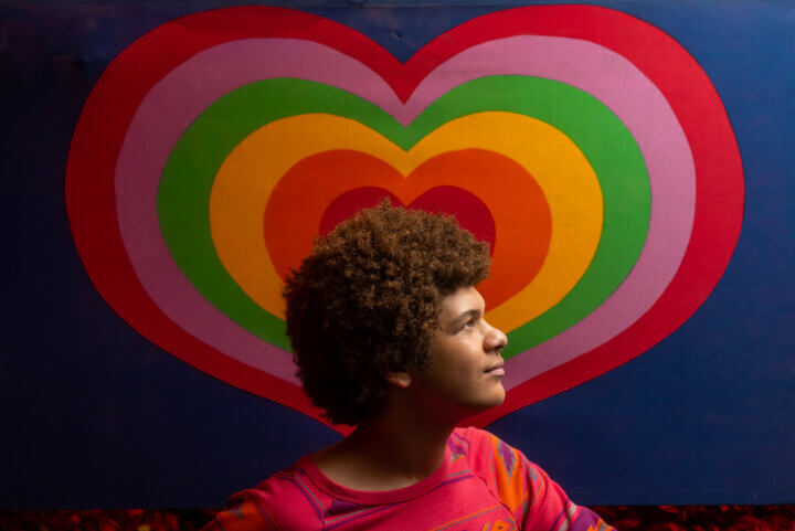 An LGBTQ+ young person stands in profile in front of a rainbow heart painting.
