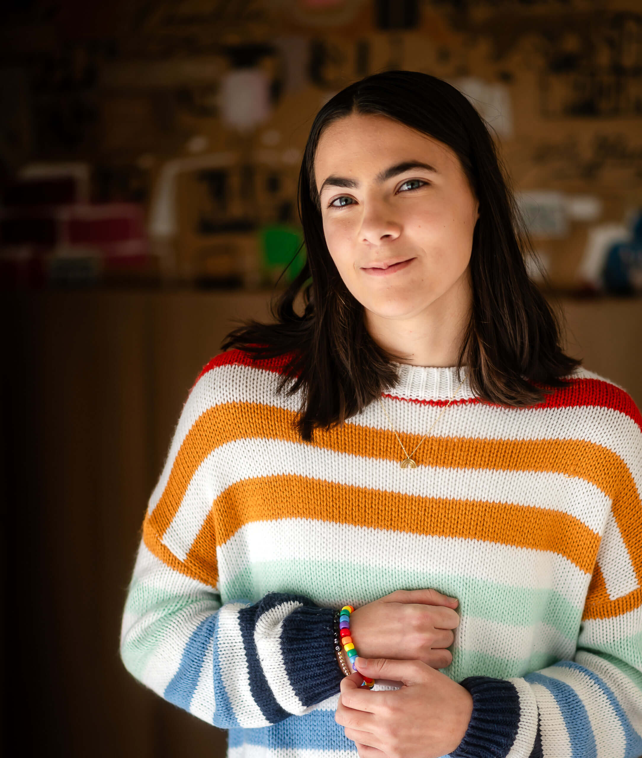 An LGBTQ+ young person in a striped sweater clutches a rainbow friendship bracelet and smiles.
