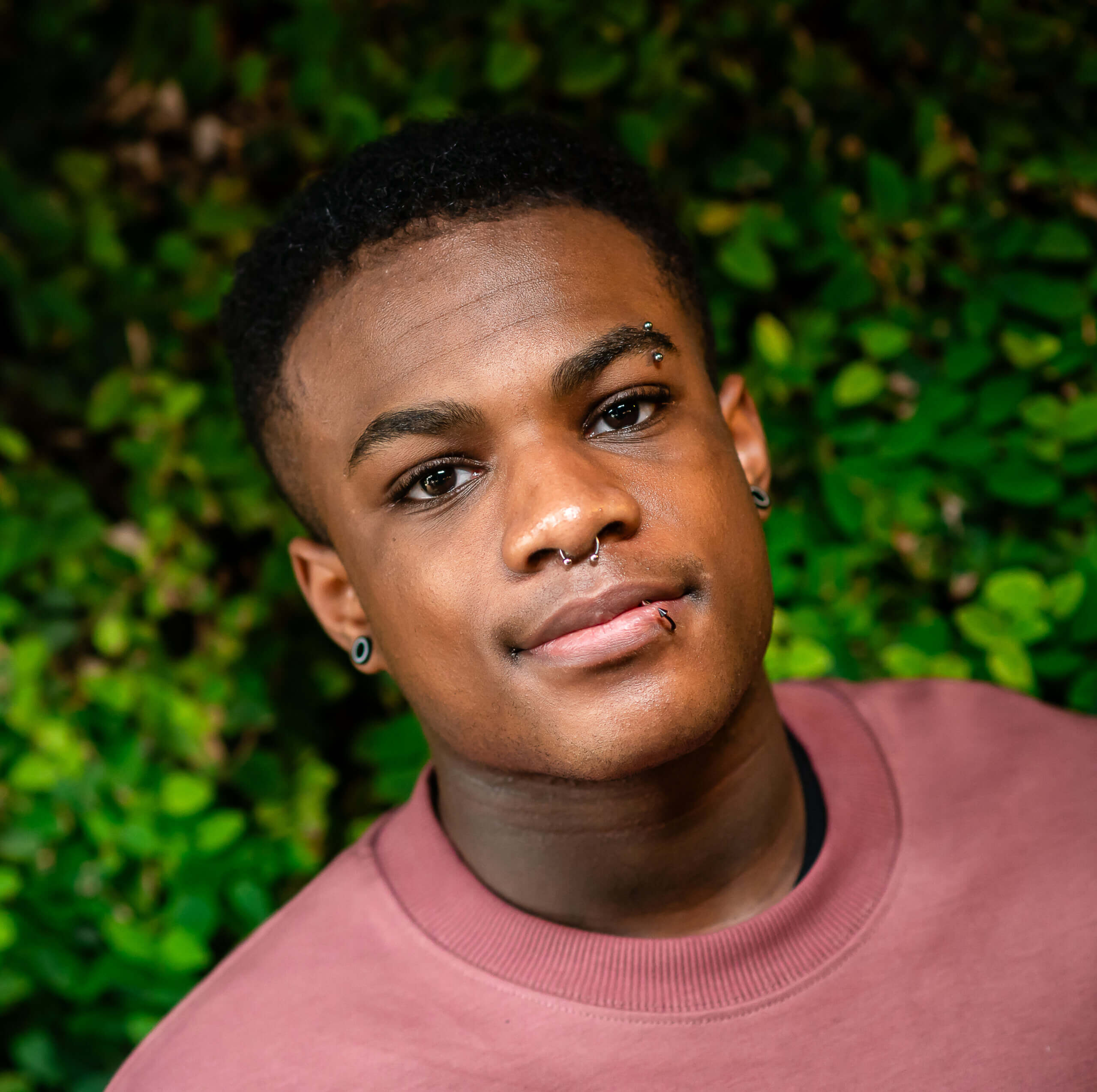 A Black LGBTQ+ young person with facial piercings smiles in front of foliage.